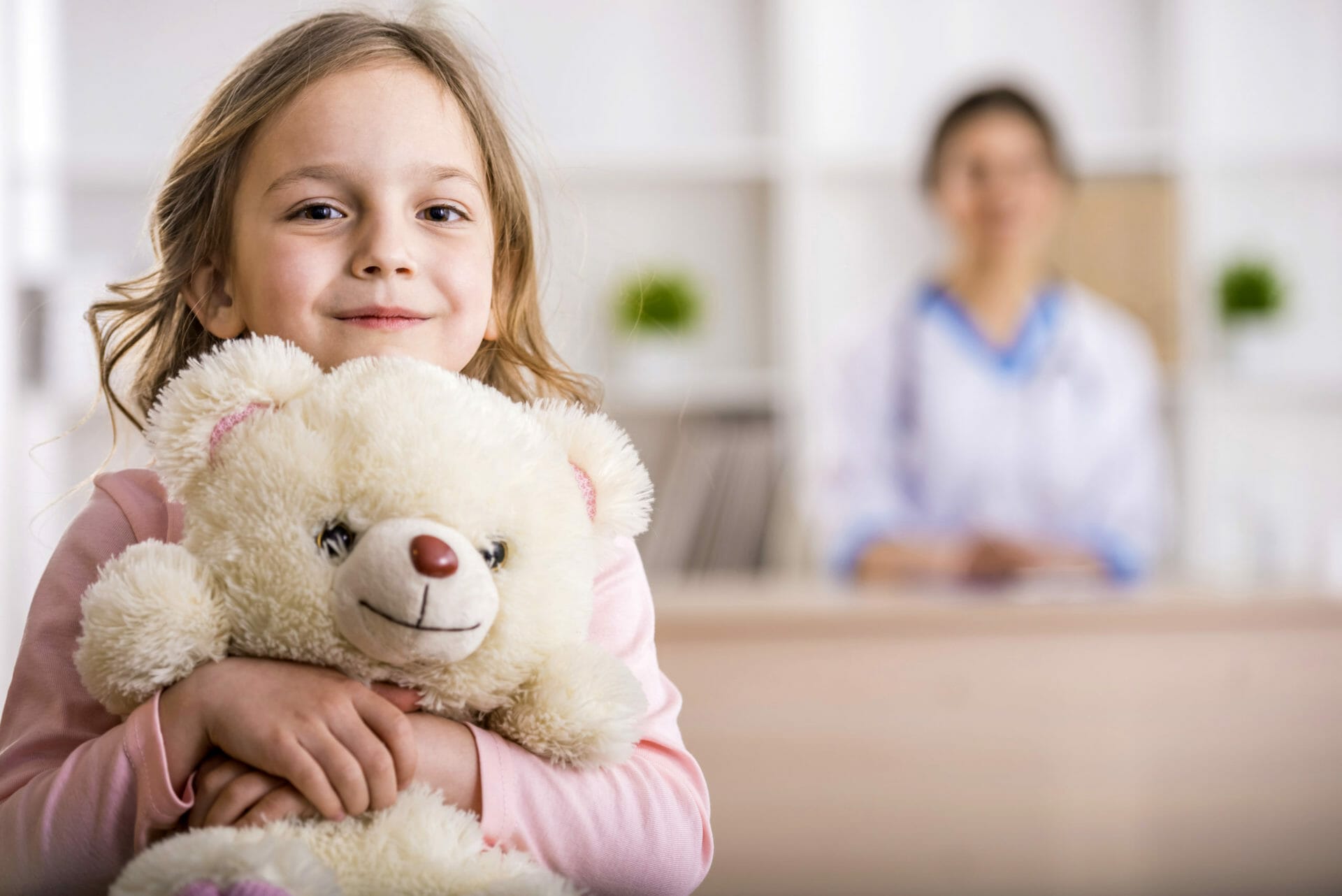 Little girl with teddy bear is looking at the camera. Female doctor on background.